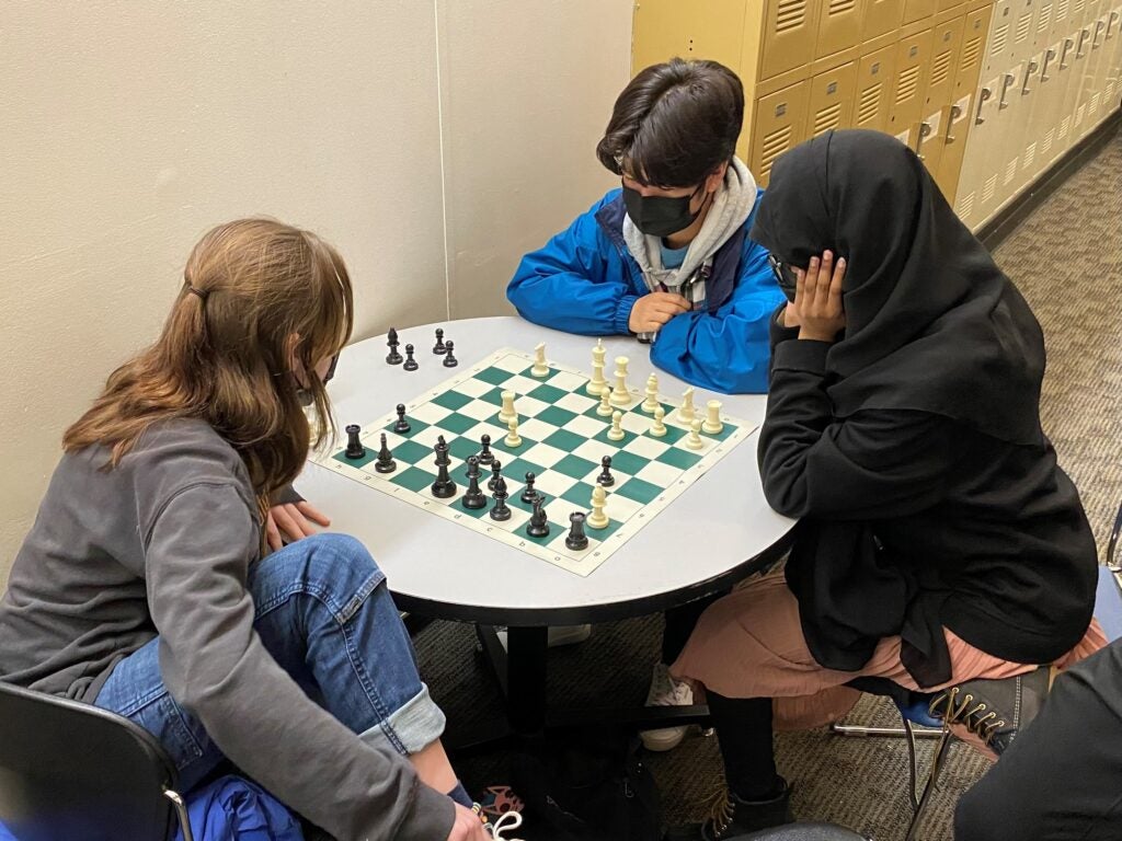3 students playing chess at round table.